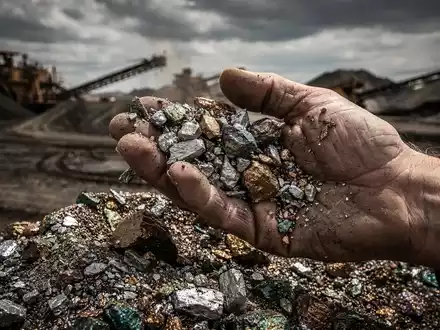 Nahaufnahme der Hand eines Bergmanns, der seltene Erden-Minerale siebt, mit verschwommenem Bergbaugebiet im Hintergrund. - Foto: über boerse-global.de