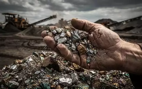 Nahaufnahme der Hand eines Bergmanns, der seltene Erden-Minerale siebt, mit verschwommenem Bergbaugebiet im Hintergrund. - Foto: über boerse-global.de