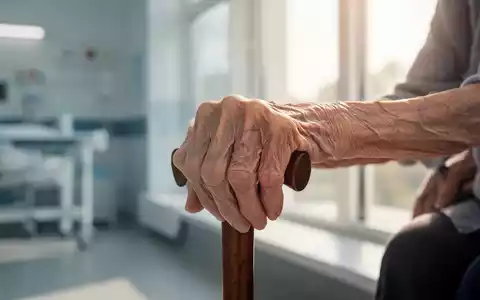 Eine alte, zerbrechliche Frauenhand ruht sanft auf einem Gehstock, im Hintergrund eine verschwommene medizinische Einrichtung. - Foto: über boerse-global.de