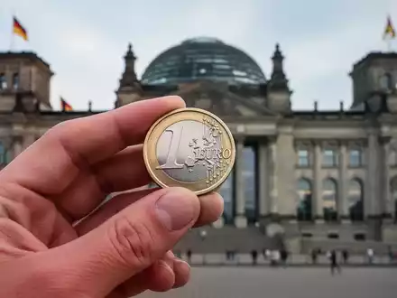 Eine deutsche Euromünze in einer Hand, im Hintergrund das verschwommene Reichstagsgebäude, symbolisiert Gesundheitsfinanzierung und Gesetzgebung. - Foto: über boerse-global.de