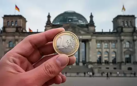 Eine deutsche Euromünze in einer Hand, im Hintergrund das verschwommene Reichstagsgebäude, symbolisiert Gesundheitsfinanzierung und Gesetzgebung. - Foto: über boerse-global.de