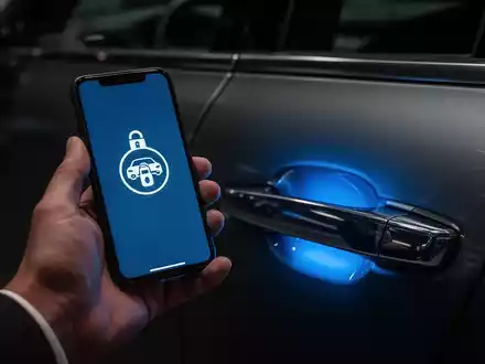 Eine Hand hält ein Smartphone, dessen Bildschirm ein Entriegelungssymbol anzeigt, das einen blauen Schein auf einen Autotürgriff wirft. - Foto: über boerse-global.de