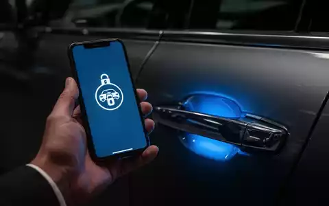 Eine Hand hält ein Smartphone, dessen Bildschirm ein Entriegelungssymbol anzeigt, das einen blauen Schein auf einen Autotürgriff wirft. - Foto: über boerse-global.de