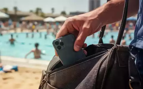 Eine Hand schiebt unauffällig ein Smartphone in eine Tasche, mit einem unscharfen, belebten Freibad oder Strand im Hintergrund. - Foto: über boerse-global.de