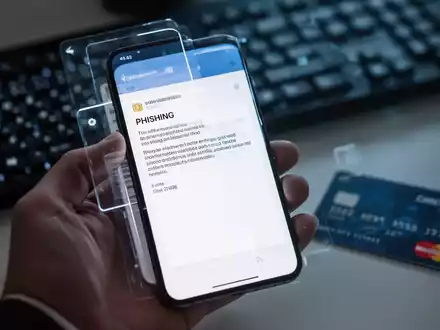 Eine Hand hält ein Smartphone mit einer Phishing-E-Mail, im Hintergrund eine Tastatur und Kreditkarte, die Cyberbetrug symbolisieren. - Foto: über boerse-global.de
