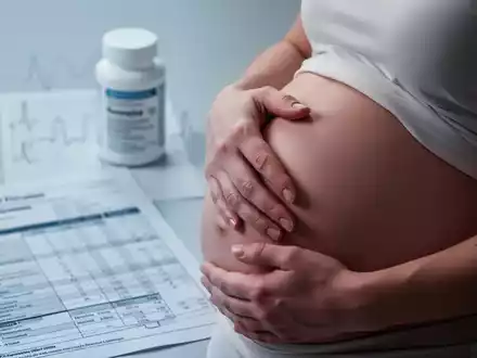 Nahaufnahme der Hand einer schwangeren Frau auf ihrem Bauch, mit verschwommenen medizinischen Elementen im Hintergrund. - Foto: über boerse-global.de
