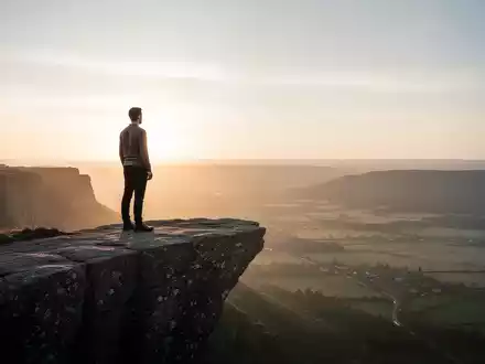 Eine Person steht auf einer Klippe und blickt auf eine weite, ruhige Landschaft bei Sonnenaufgang, die Widerstandsfähigkeit symbolisiert. - Foto: über boerse-global.de