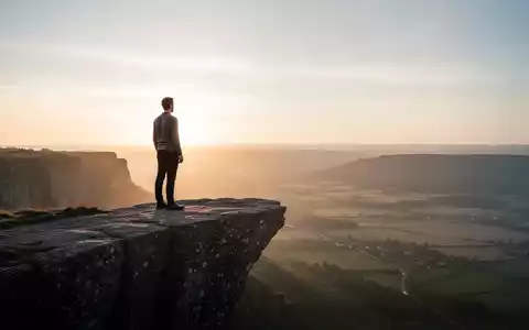 Eine Person steht auf einer Klippe und blickt auf eine weite, ruhige Landschaft bei Sonnenaufgang, die Widerstandsfähigkeit symbolisiert. - Foto: über boerse-global.de