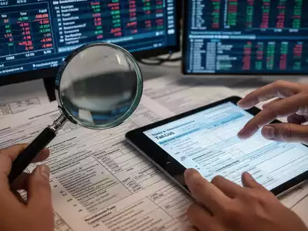Eine Lupe vergrößert einen Abschnitt komplexer Steuerdokumente, während im Hintergrund digitale Bildschirme Finanzdaten anzeigen. - Foto: über boerse-global.de