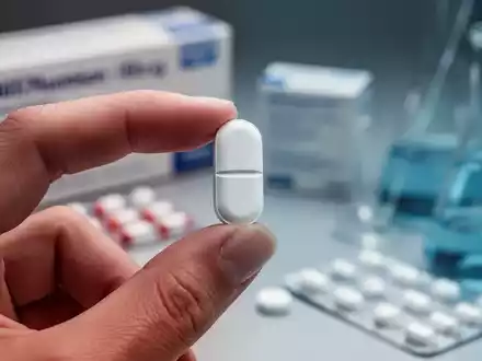 Eine Person hält eine weiße Pille zwischen Daumen und Zeigefinger, im Hintergrund verschwommene pharmazeutische Verpackungen und Instrumente. - Foto: über boerse-global.de