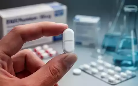 Eine Person hält eine weiße Pille zwischen Daumen und Zeigefinger, im Hintergrund verschwommene pharmazeutische Verpackungen und Instrumente. - Foto: über boerse-global.de