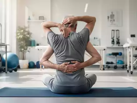 Eine ältere Person, von hinten gesehen, führt eine sanfte Wirbelsäulenbeugeübung auf einer Yogamatte in einem Therapieraum durch. - Foto: über boerse-global.de