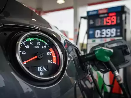 Nahaufnahme einer Tankanzeige, die einen vollen Tank anzeigt, mit einer unscharfen Tankstelle im Hintergrund. - Foto: über boerse-global.de