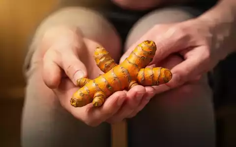 Eine Hand hält eine leuchtend gelbe Kurkumawurzel, im verschwommenen Hintergrund sind Knie angedeutet. - Foto: über boerse-global.de