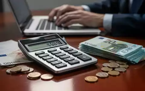 Ein Nahaufnahme-Bild eines Taschenrechners mit Euro-Banknoten und Münzen auf einem Schreibtisch, im Hintergrund tippt eine Person auf einer Laptoptastatur. - Foto: über boerse-global.de