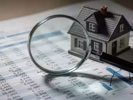 Eine Lupe vergrößert einen Finanzbericht mit Zahlen und Diagrammen, hinter der ein kleines Hausmodell steht, um die Prüfung von Immobilienwerten darzustellen. - Foto: über boerse-global.de
