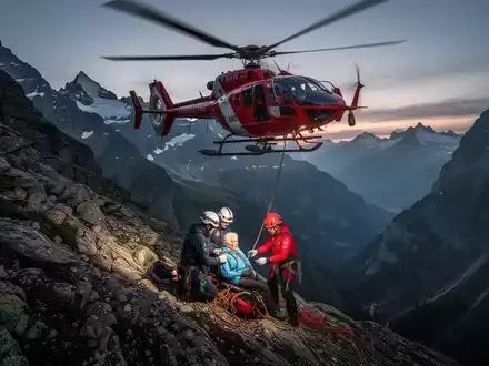 Rettungshubschrauber über steilem Schweizer Berghang bei Sonnenuntergang, Rettungskräfte kümmern sich um eine ältere Person. - Foto: über boerse-global.de