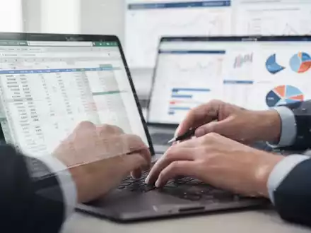 Nahaufnahme von Händen, die an einer Laptop-Tabelle arbeiten, mit verschwommenen Finanzdaten im Hintergrund, symbolisiert Excel-Kenntnisse. - Foto: über boerse-global.de