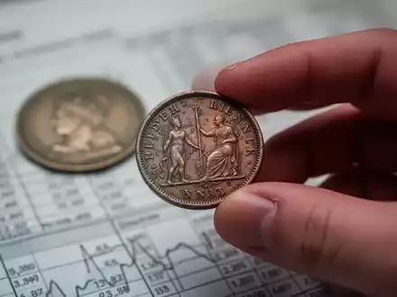 Nahaufnahme einer Hand, die eine britische Penny-Münze hält, mit einer weiteren Münze im Hintergrund, die kleine finanzielle Differenzen symbolisiert. - Foto: über boerse-global.de