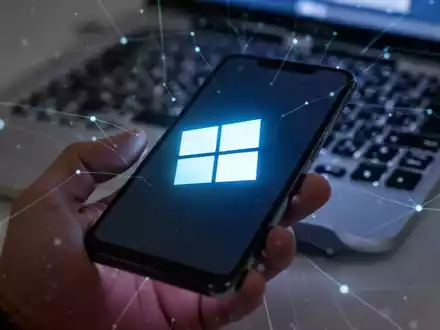 Ein stilisiertes Windows 11-Logo auf einem Smartphone-Bildschirm, das in einer Hand gehalten wird, im Hintergrund eine verschwommene Laptop-Tastatur. - Foto: über boerse-global.de