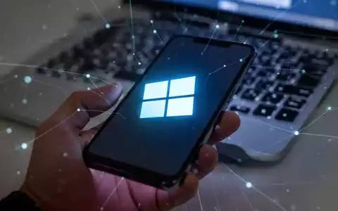 Ein stilisiertes Windows 11-Logo auf einem Smartphone-Bildschirm, das in einer Hand gehalten wird, im Hintergrund eine verschwommene Laptop-Tastatur. - Foto: über boerse-global.de