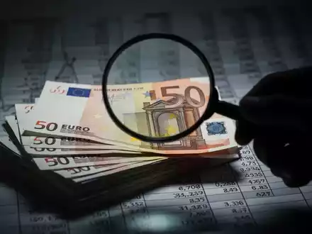 Ein Nahaufnahme von Euro-Banknoten, teilweise verdeckt von einer Hand mit einer Lupe, die finanzielle Prüfung und Untersuchung symbolisiert. - Foto: über boerse-global.de