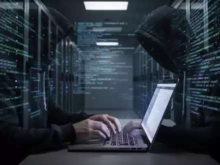 Eine Person im Kapuzenpullover tippt an einem Laptop in einem dunklen Serverraum, mit digitalem Code an den Wänden. Fokus auf Hände und Bildschirm. - Foto: über boerse-global.de