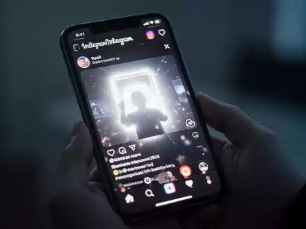 Nahaufnahme eines iPhone-Bildschirms mit Instagram Reels und hellem, überbelichtetem Glühen, gehalten in einer Hand in einem schwach beleuchteten Raum. - Foto: über boerse-global.de