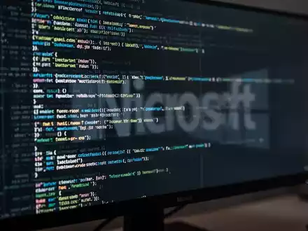 Stilisierte Nahaufnahme eines Computermonitors mit abstraktem Code und CLI-Elementen, dezente Microsoft-Logo-Reflexion. - Foto: über boerse-global.de