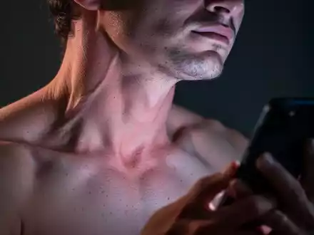 Nahaufnahme einer Person mit sichtbaren Nacken- und Schulterverspannungen, beleuchtet vom Schein eines Smartphones. - Foto: über boerse-global.de