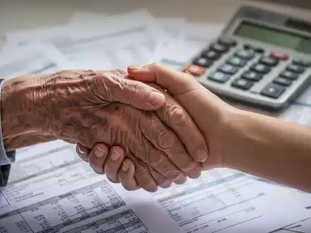 Eine ältere Hand hält sanft eine jüngere Hand, im Hintergrund verschwommen Finanzdokumente und ein Taschenrechner. - Foto: über boerse-global.de