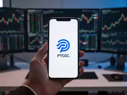 Eine Hand hält ein Smartphone mit Stablecoin-Logo vor einem unscharfen Handelstisch mit mehreren Monitoren. - Foto: über boerse-global.de