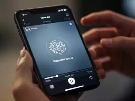 Nahaufnahme eines iPhone-Bildschirms mit eingefrorenem Video und unscharfen Händen im Hintergrund, symbolisiert Wiedergabeprobleme. - Foto: über boerse-global.de