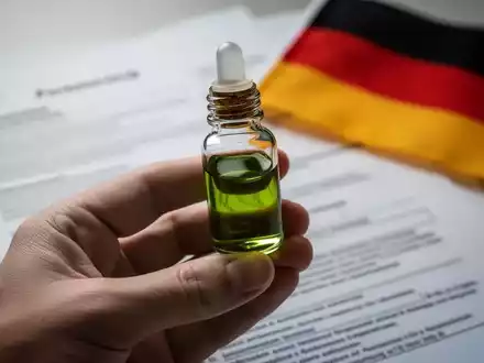 Eine Hand hält eine kleine Flasche medizinisches Cannabisöl, im Hintergrund verschwommen juristische Dokumente und eine deutsche Flagge. - Foto: über boerse-global.de