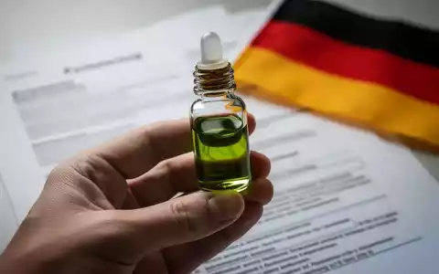 Eine Hand hält eine kleine Flasche medizinisches Cannabisöl, im Hintergrund verschwommen juristische Dokumente und eine deutsche Flagge. - Foto: über boerse-global.de