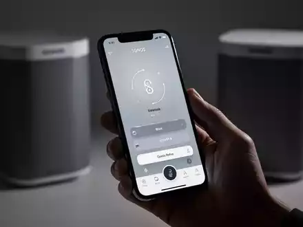 Eine Hand hält ein iPhone mit der neuen Sonos-App-Oberfläche, tab-basierter Navigation und Lautstärkereglern, im Hintergrund ein unscharfer Sonos-Lautsprecher. - Foto: über boerse-global.de