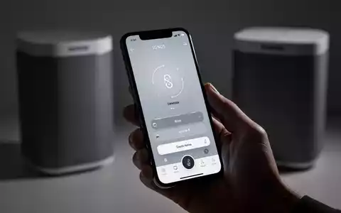 Eine Hand hält ein iPhone mit der neuen Sonos-App-Oberfläche, tab-basierter Navigation und Lautstärkereglern, im Hintergrund ein unscharfer Sonos-Lautsprecher. - Foto: über boerse-global.de
