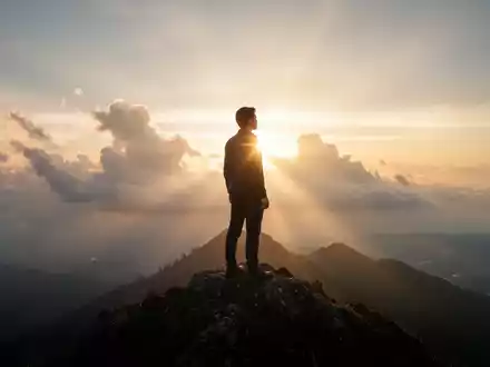 Eine Person steht bei Sonnenaufgang auf einem Berggipfel und blickt über eine weite, neblige Landschaft. - Foto: über boerse-global.de