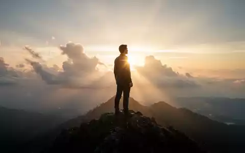 Eine Person steht bei Sonnenaufgang auf einem Berggipfel und blickt über eine weite, neblige Landschaft. - Foto: über boerse-global.de