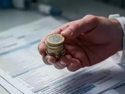 Eine Hand hält eine kleine Ansammlung von Euro-Münzen, im unscharfen Hintergrund sind medizinische Dokumente zu erkennen. - Foto: über boerse-global.de