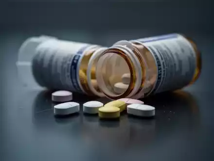 Eine Nahaufnahme zeigt eine unscharfe Pillendose und lose Tabletten auf einer dunklen, spiegelnden Oberfläche. - Foto: über boerse-global.de
