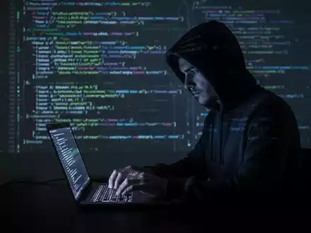 Verschattete Gestalt in Kapuzenjacke tippt auf Laptop, dahinter Code-Zeilen projiziert, symbolisiert Cyberkriminalität und Datendiebstahl. - Foto: über boerse-global.de
