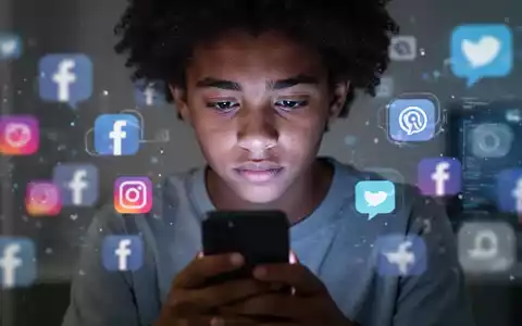 Ein nachdenklicher Teenager schaut auf ein Smartphone, das Gesicht vom Bildschirmlicht beleuchtet, umgeben von verschwommenen digitalen Symbolen. - Foto: über boerse-global.de
