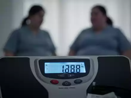Eine Nahaufnahme einer digitalen Waage, die ein hohes Gewicht anzeigt, mit einer unscharfen Silhouette einer Person im Hintergrund. - Foto: über boerse-global.de