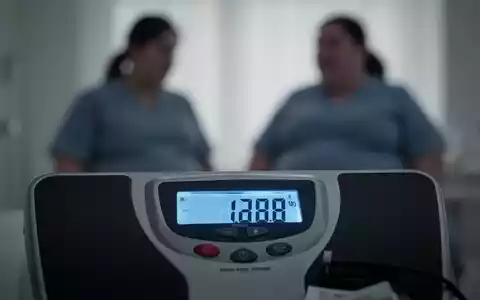 Eine Nahaufnahme einer digitalen Waage, die ein hohes Gewicht anzeigt, mit einer unscharfen Silhouette einer Person im Hintergrund. - Foto: über boerse-global.de