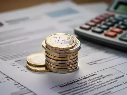 Nahaufnahme eines Stapels deutscher Euro-Münzen mit unscharfem Hintergrund aus offiziellen Dokumenten und einem Taschenrechner. - Foto: über boerse-global.de