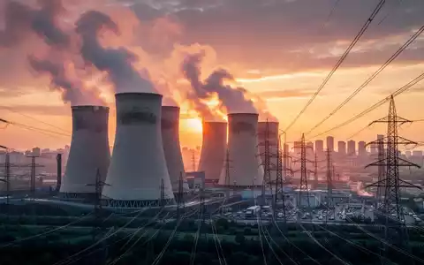 Ein großes Kohlekraftwerk mit Kühltürmen bei Sonnenuntergang, Hochspannungsleitungen führen zu einer Stadt. - Foto: über boerse-global.de