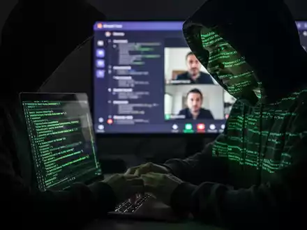 Verschwommene Gestalt in Kapuzenjacke tippt auf Laptop, im Hintergrund unscharfer Microsoft-Teams-Anruf, symbolisiert Vishing-Angriff. - Foto: über boerse-global.de