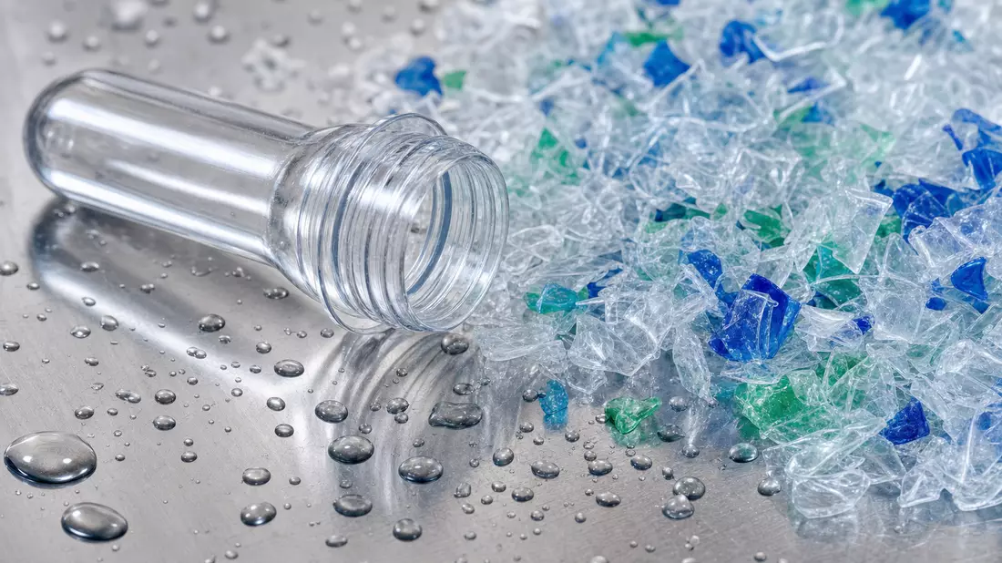 Makroaufnahme eines PET-Kunststoff-Preforms und farbiger Recycling-Flakes auf glänzender Metalloberfläche