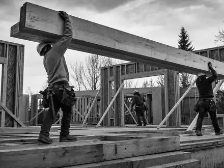 Builders FirstSource US12189T1043 ZimmermĂ€nner tragen schwere Holzbalken auf einer Wohnhaus-Baustelle im SchwarzweiĂfoto - Foto: THN